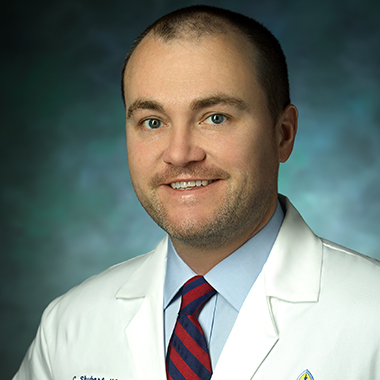 Chris Shubert, in a formal portrait, wearing a white lab coat, light blue button down shirt and red and blue striped tie