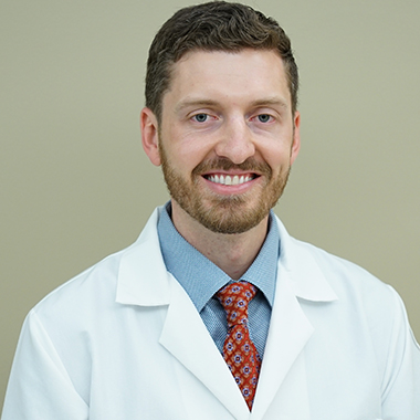 Brett Parker, in a formal portrait, wearing a white lab coat, blue and white button down shirt and red tie