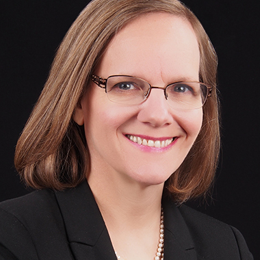 Jessica Bienstock, in a formal portrait, wearing a black blazer and pearl necklace