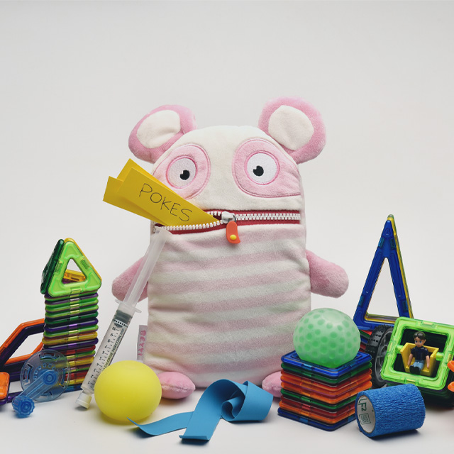 A group of toys and medical supplies, including a syringe and medical tape. In the center is a pink plush panda, with slips of paper protruding from the zippered pocket of its mouth. One of the slips says 'Pokes'.