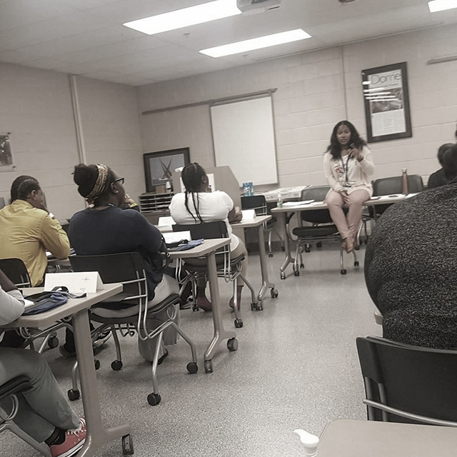 A black and white photo of students sit in a classroom while listening to a women at the front.