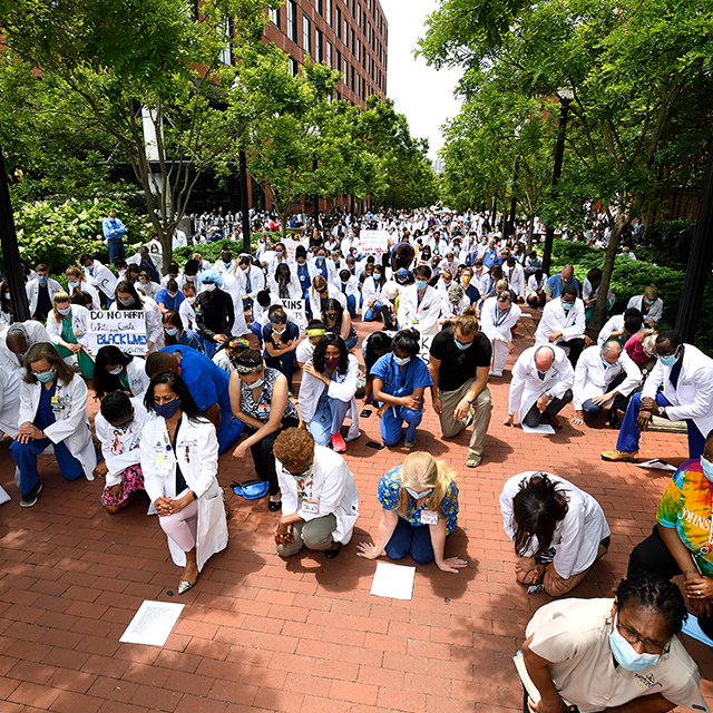 Hundreds of Johns Hopkins clinicians in white lab coats kneel on the campus of the Johns Hopkins hospital.