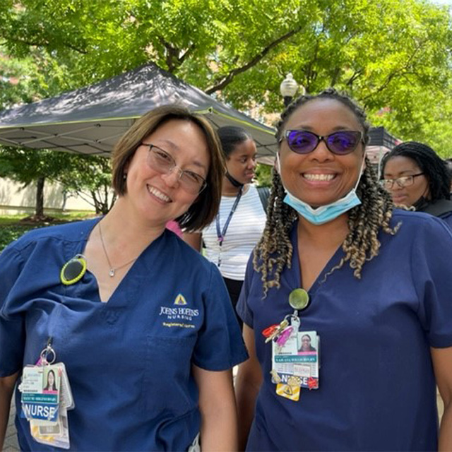 Nurses Mayumi Shigeno and Lajuana Willis, wearing blue scrubs and smiling, with the tents of the farmers’ market behind them. 