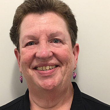 A headshot of Melissa Gerstenhaber wearing a black polo shirt, gold necklace and purple earrings