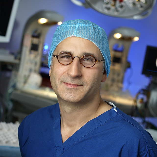 Ahmet Baschat poses in the operating room wearing blue scrubs and a surgical cap, with equipment behind him