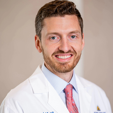 Brett Parker, in a formal portrait, wearing a white lab coat, light blue button down shirt and red tie.