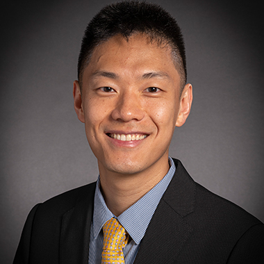 Risheng Xu, in a formal portrait, wearing a black suit and light blue striped button down shirt with a yellow tie