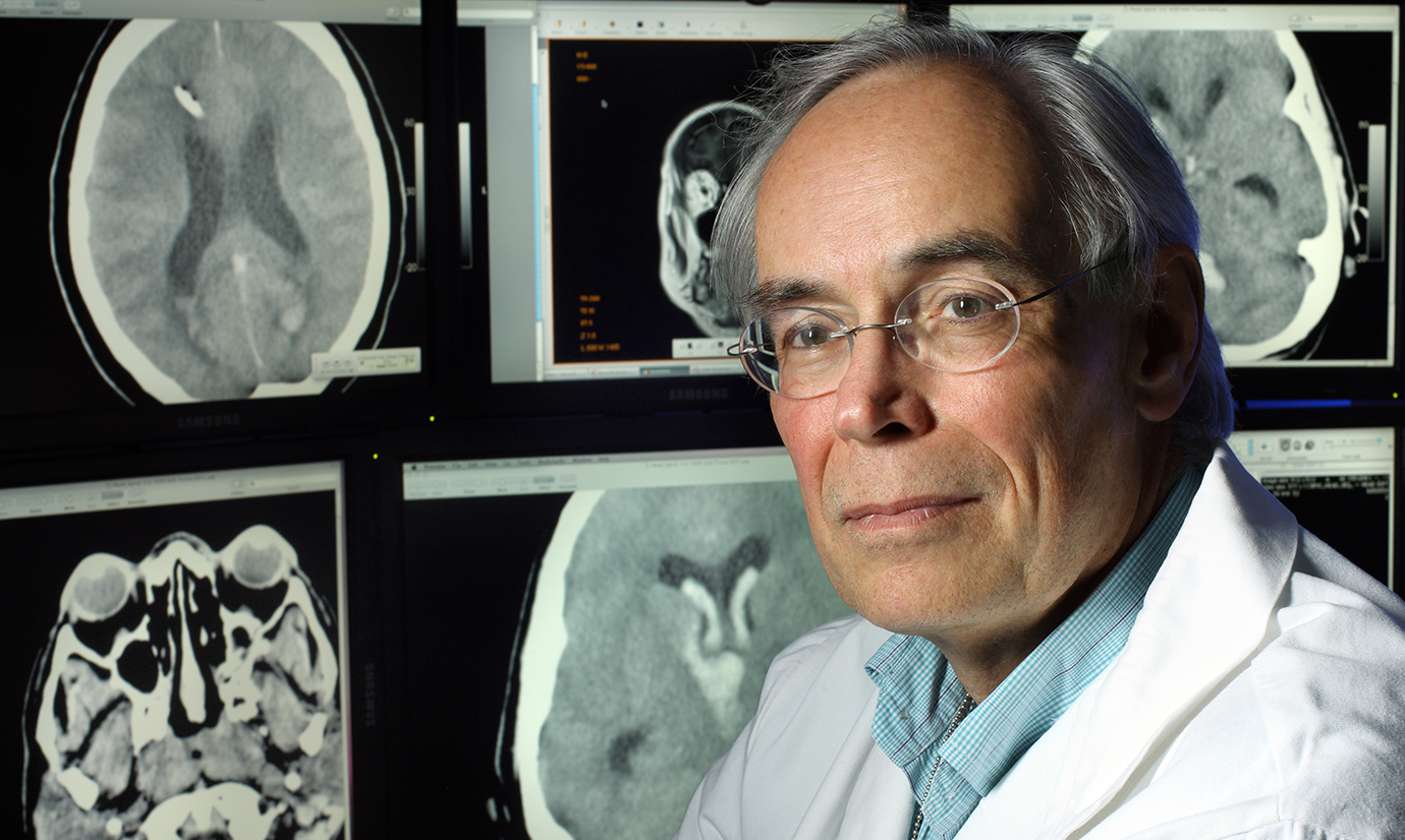 Daniel Hanley, wearing a white lab coat and light blue button down shirt, poses in front of computer monitors showing brain imaging