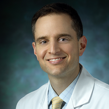 Head shot of a smiling Dr. Hasan, wearing a white coat over a blue shirt and yellow tie. 