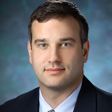 Eric Jackson, in a formal portrait, wearing a dark blue suit, light blue button down shirt and blue, black and white striped tie
