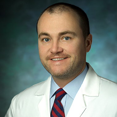 Surgical oncologist Christopher Shubert in a formal portrait, wearing a white lab coat, light blue button down and red and blue striped tie.