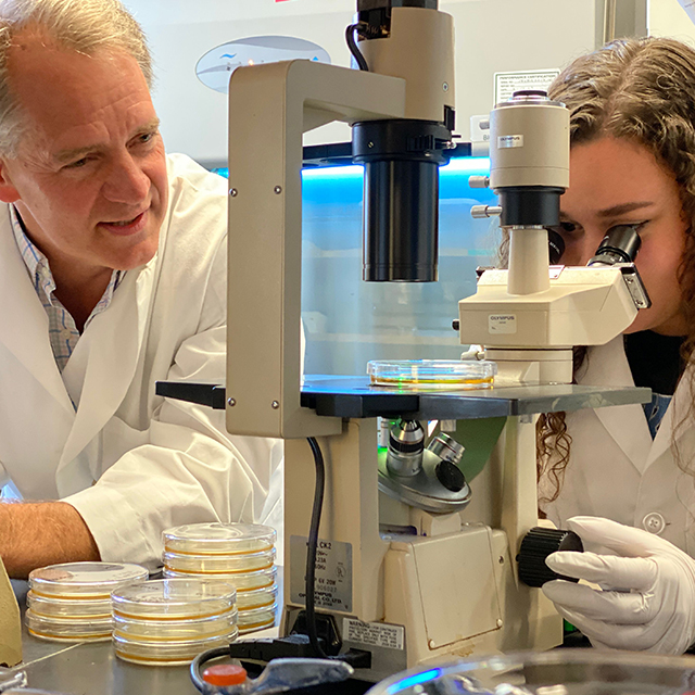 Doug Robinson looks on as Eleana Parajón, wearing white gloves, peers into a microscope. Both are wearing white coats. 
