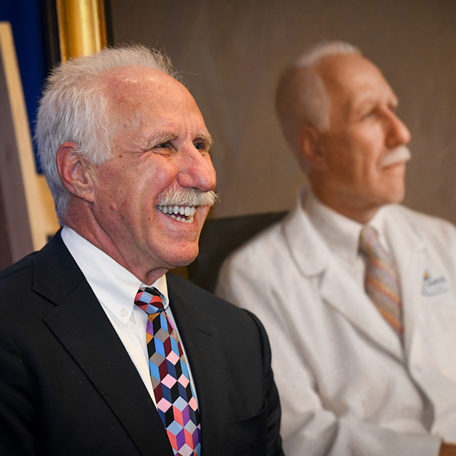 A laughing Rothman, in dark jacket and bright tie, in the foreground, with his portrait behind him.