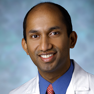 Chetan Bettegowda, in a formal portrait, wearing a white lab coat, red tie and blue button-down shirt.