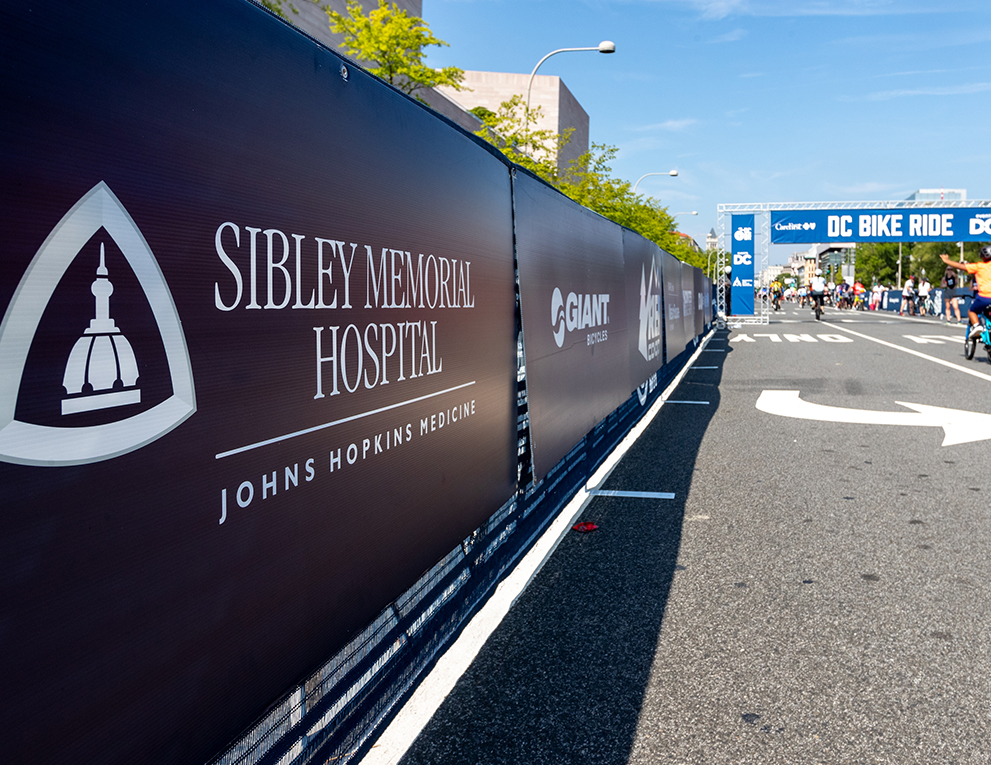 A black poster mounted to a fence reads "Sibley Memorial Hospital: Johns Hopkins Medicine."