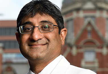 Shah stands in front of the billings building, wearing a white coat. 