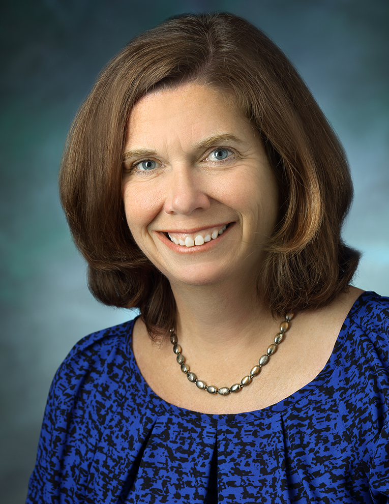 Sue De Pasquale smiles and stands against a blue-grey background.