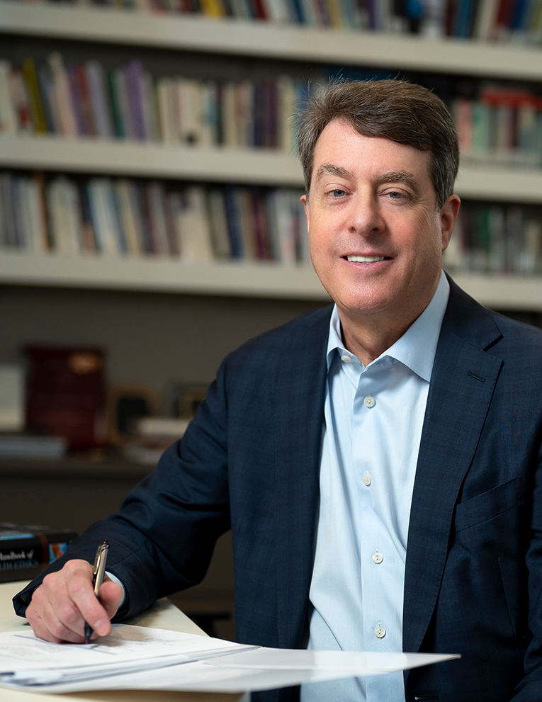 Jeffrey Khan sits at a desk, pen in hand, as he smiles at the camera.