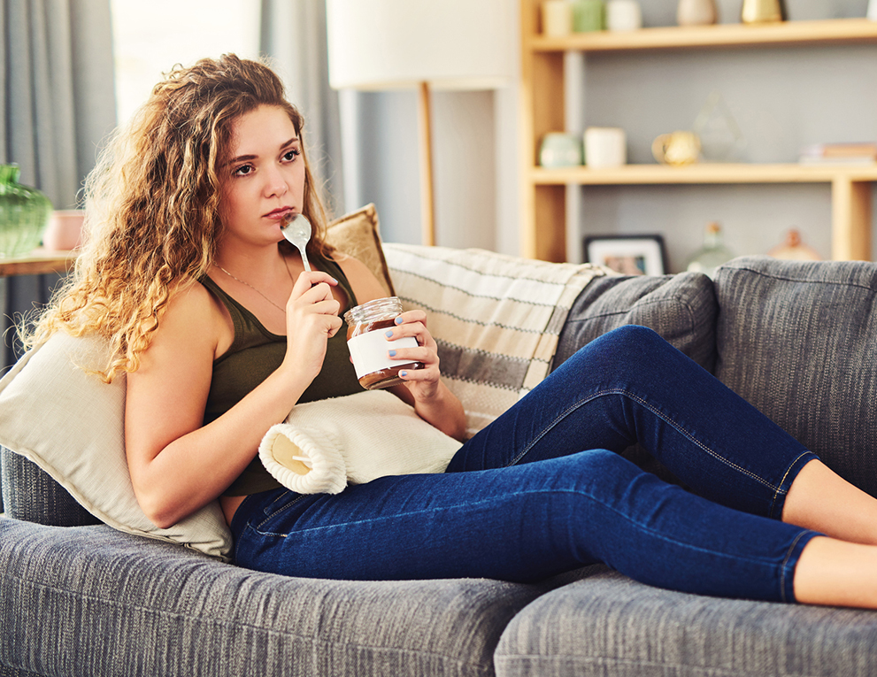 A woman sits on her couch with a jar of peanut butter, a spoon and a heating pad.