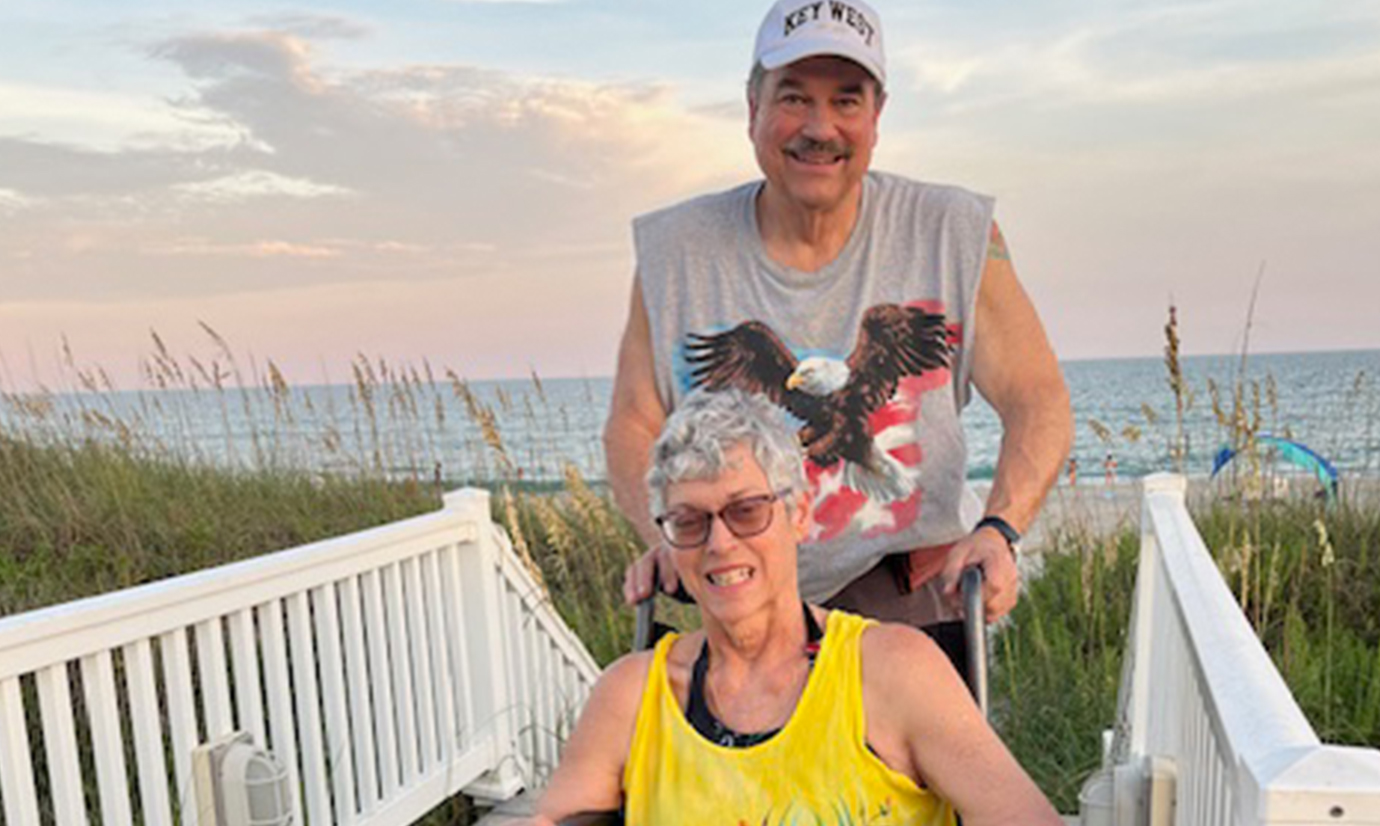 A husband stands behind his wife who sits in a wheelchair. 