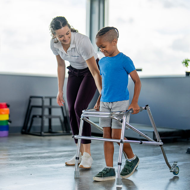A rehabilitation expert helping a young child walk with the assistance of a walker.