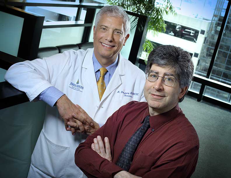 Two doctors stand next to each other in an office setting, bright window behind them, both smiling.