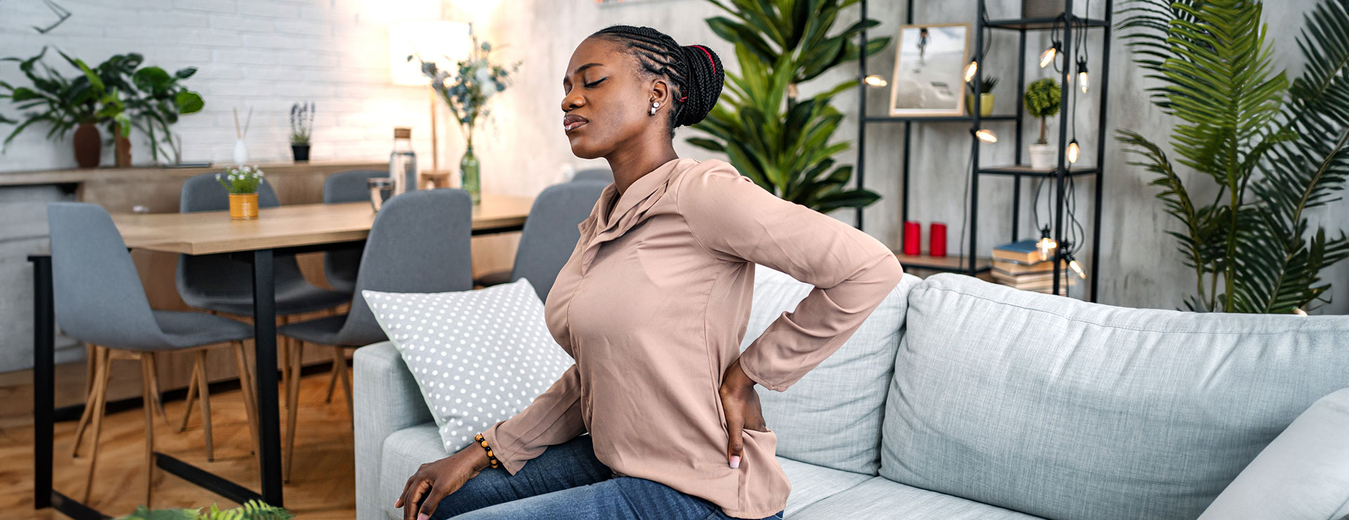 A woman sitting on her couch, holding her back in pain.