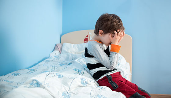 A small boy holds his head in his hands while sitting on the edge of his bed.