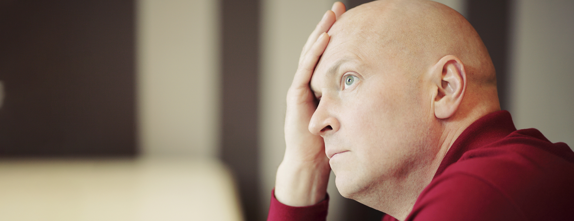 A bald man in a red shirt holds his hand to his forehead, looking preoccupied.