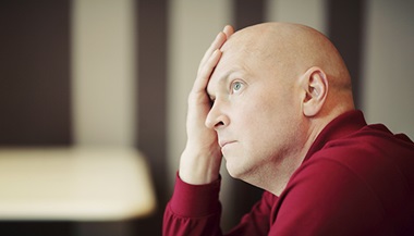 A bald man in a red shirt holds his hand to his forehead, looking preoccupied.