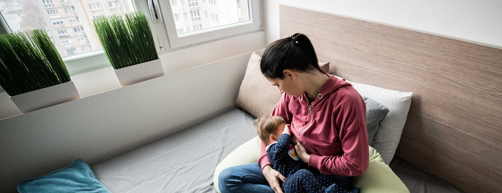 A woman sits on her bed, nursing her baby.