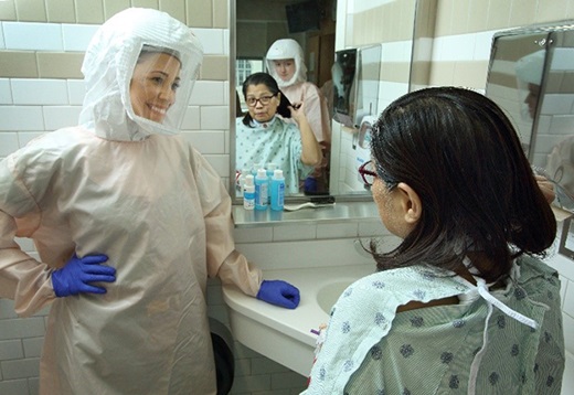 An occupational therapist wearing PPE looks on as a patient brushes her hair.