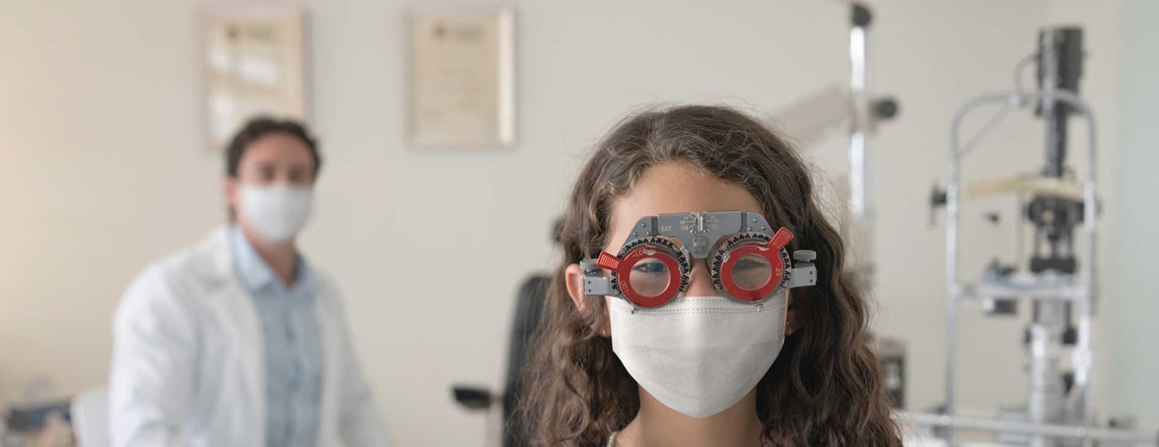 A young girl getting an eye exam with an eye doctor in the background.