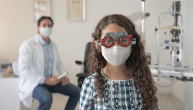A young girl getting an eye exam with an eye doctor in the background.