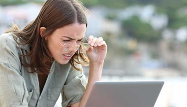 A young woman squints while lifting her glasses to see her laptop screen