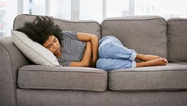 A woman lays on the couch, hugging her stomach in pain.