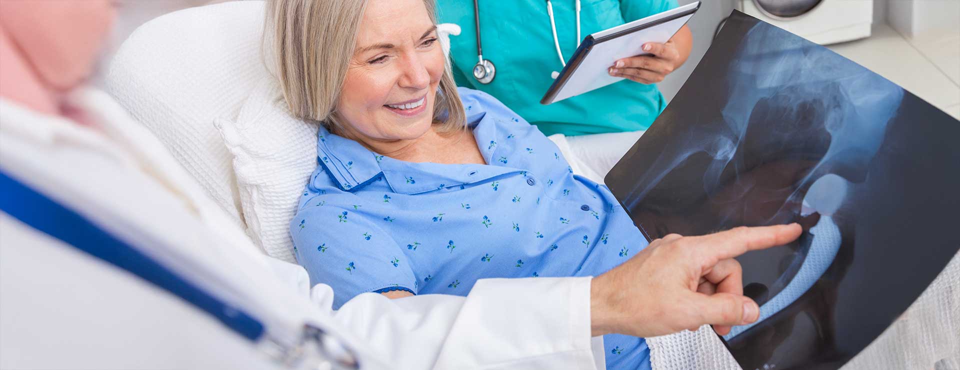 A smiling older woman in a hospital bed looks at a hip X-ray with a doctor, who is pointing at it.. A nurse stands nearby holding a tablet.