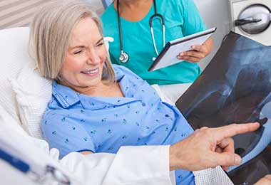 A smiling older woman in a hospital bed looks at a hip X-ray with a doctor, who is pointing at it.. A nurse stands nearby holding a tablet.