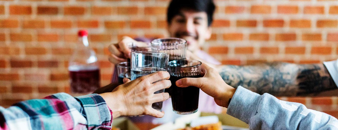 A group of friends clink their glasses of soda together over pizza.