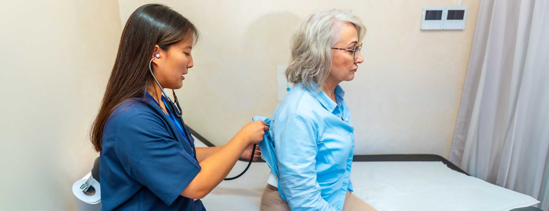 A nurse in blue uses a stethoscope to check an older woman in a clinic during a lung exam; both look focused and calm.