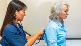 A nurse in blue uses a stethoscope to check an older woman in a clinic during a lung exam; both look focused and calm.