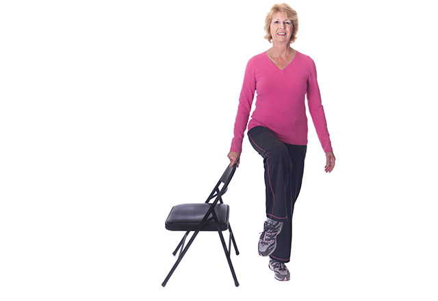 A senior woman exercises using a chair for support.