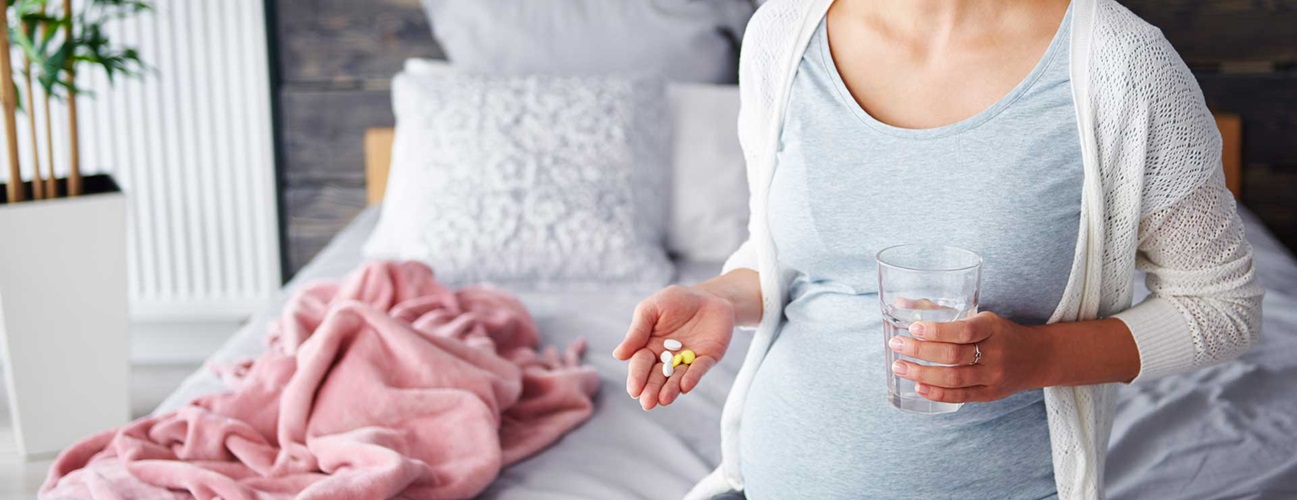 A pregnant woman sits on the edge of her bed, holding a glass of water and medication.