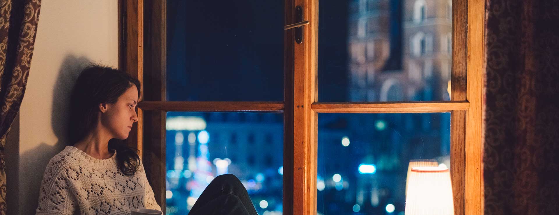 A woman looks outside her window with a sad expression.