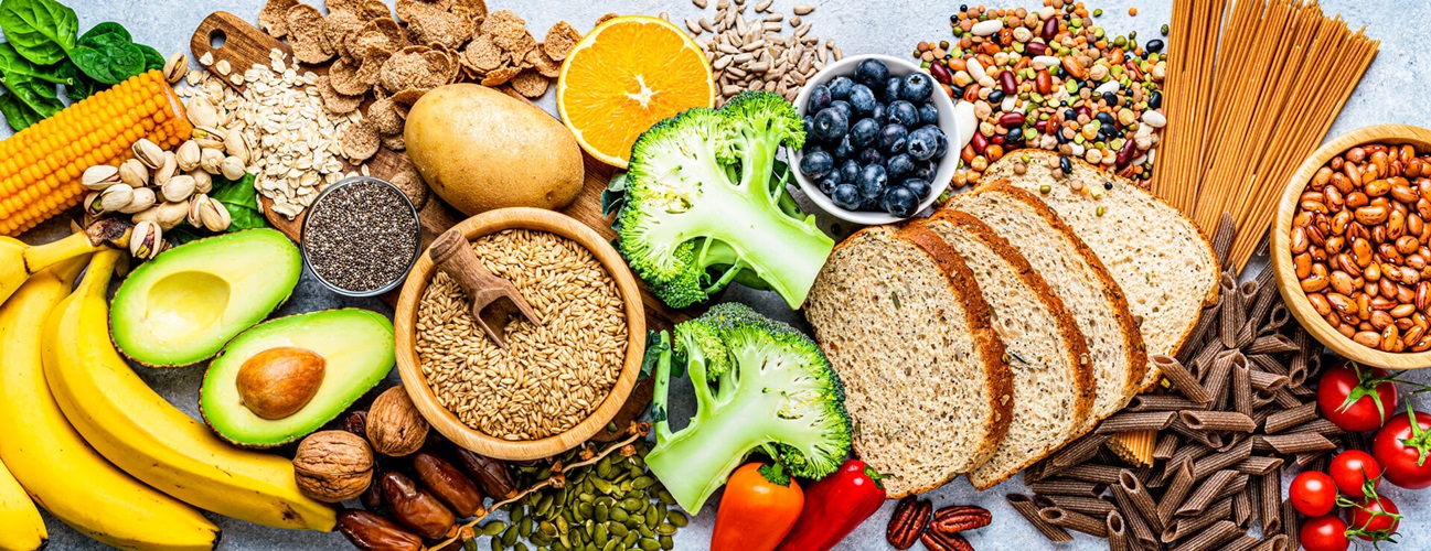 Overhead view of a large group of food with high content of dietary fiber arranged side by side. The composition includes berries, oranges, avocado, chia seeds, wholegrain bread, wholegrain pasta, whole wheat, potatoes, oat, corn, mixed beans, brazil nut, sunflower seeds, pumpkin seeds, broccoli, pistachio, banana among others. 