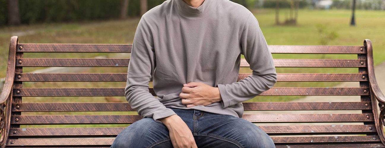 A young man sits on a bench outside, holding his stomach in pain.