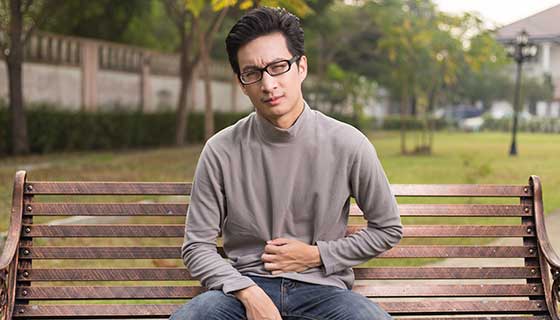 A young man sits on a bench outside, holding his stomach in pain.