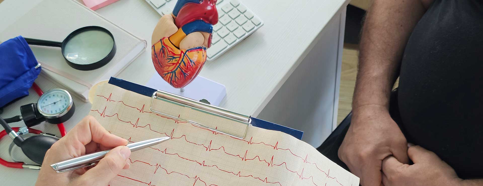 A doctor points at a heart rate graph on a clipboard with a pen, while a patient listens attentively. A heart model and medical tools are on the desk.