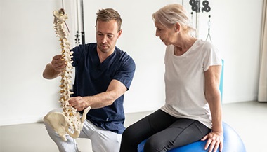 A clinician showing a patient a location on a model of a spine while they sit on balancing balls.