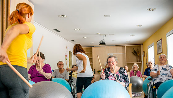 A recreational therapist working with a class of seniors with drum sticks and balancing balls.